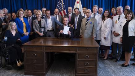 Governor Newsom sitting at his desk and holding up the signed AB 35 bill surrounded by attendees
